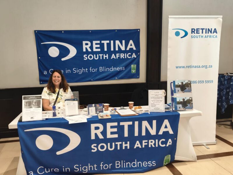 The depicts a promotional booth for “Retina South Africa,” at the Transport Summit. The booth features a blue banner with the Retina SA logo and name, the tagline “A Cure in Sight for Blindness,” seated at the booth is Mariza Jurgens (Vice Chairperson) of Retina SA.