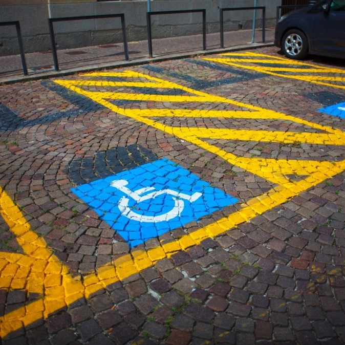 A cobblestone pavement with a painted symbol indicating a disabled parking space. The symbol is a white pictogram of a person in a wheelchair set against a blue square background. Surrounding this blue square is a yellow hatched pattern, suggesting no parking or restricted access, typically used to denote the boundaries of the reserved space.