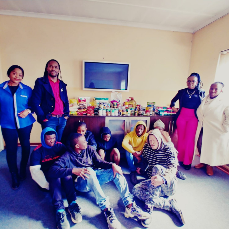 A groupd of individuals from the Johannesburg Society for the Blind and the two Retina Social Workers behind them displayed on a table is all the food items that were donated for Mandela day.
