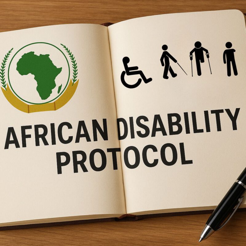 An open notebook on a wooden surface displays the title "African Disability Protocol" in bold black letters. On the left page is a green silhouette of the African continent encircled by a golden emblem with laurel leaves, and on the right is a black disability icons. A black pen lies beside the notebook
