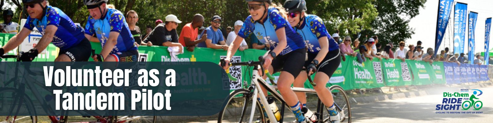 Group of cyclists riding tandem bicycles during a public event. Sighted pilots lead visually impaired riders, all wearing matching blue jerseys and helmets. Spectators cheer from behind green barriers. Event signage promotes volunteering as a tandem pilot and cycling to restore sight
