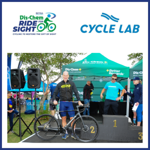 Jason stands on a stage holding the Orbea bicycle in front of a podium setup at an outdoor event. Behind the cyclist are green tents displaying the Dis‑Chem branding and event staff standing near the podium steps. Large speakers are positioned on the left side. At the top of the image, two logos appear: “Dis‑Chem Ride for Sight – Cycling to Restore the Gift of Sight” on the left and “Cycle Lab” on the right.
