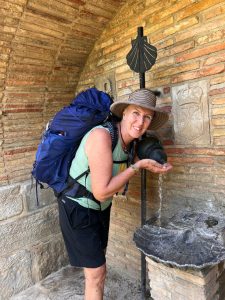Carine with a large backpack drinking water from a wall-mounted fountain along the Camino route, standing beside a brick wall with a scallop shell symbol above the fountain.
