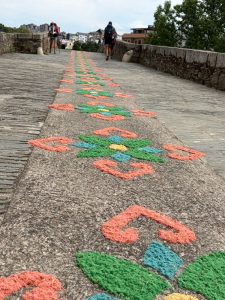 Image (Image_23-SB.jpg — Corpus Christi alfombra on the bridge) "A close-up view looking along a stone bridge in Ponferrada, Spain, where an intricate traditional alfombra — a decorative carpet made from dyed sawdust and sand — stretches the full length of the bridge in vibrant orange, green, teal and yellow patterns. Two pilgrims with backpacks walk in the distance. The alfombra was created for the Octava de Corpus Christi procession 2026.