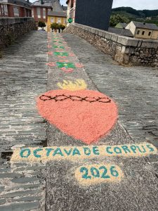 A close-up view of a traditional alfombra on a stone bridge in Ponferrada, Spain, created for the Corpus Christi procession. In the foreground, a large pink Sacred Heart with a crown of thorns and yellow flames is depicted in dyed sawdust and sand, with the words 'Octava de Corpus 2026' written below in teal lettering on a cream background. The decorative carpet of colourful geometric patterns continues along the length of the bridge into the distance.