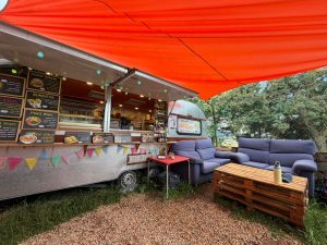 The exterior of El Paraiso, a converted silver caravan café on the Camino de Santiago, sheltered under a large orange awning. Chalkboard menus on the side of the caravan advertise veggie burgers, burritos, Thai poke bowl, hot spicy lentil soup, tomato avocado toast, hummus toast and guacamole toast. Two blue sofas and a wooden pallet coffee table are arranged on the gravel outside, creating a relaxed seating area. The El Paraiso logo is visible on the side of the caravan, with trees in the background.