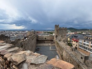 View from the battlements of the Castillo de los Templarios in Ponferrada, Spain, looking down into the open stone courtyard below. The castle's crenellated towers rise on the right, with the rooftops and buildings of Ponferrada city spreading out beyond the walls beneath a dramatic overcast sky.
