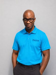 Joseph wearing glasses and a bright blue Retina South Africa polo shirt, photographed against a plain grey studio background.