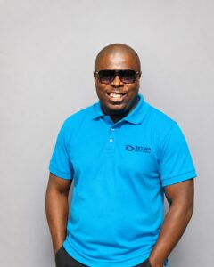 Unarine smiling wearing sunglasses and a bright blue Retina South Africa polo shirt, photographed against a plain grey studio background