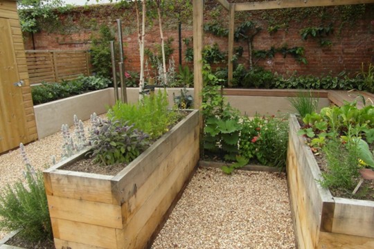 A walled garden with raised wooden planter beds filled with herbs and leafy plants. Gravel paths run between the beds, and a red brick wall covered in climbing plants stands at the back.